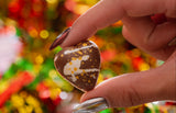 Hand holding a heart-shaped chocolate against a festive, blurred background with lights.